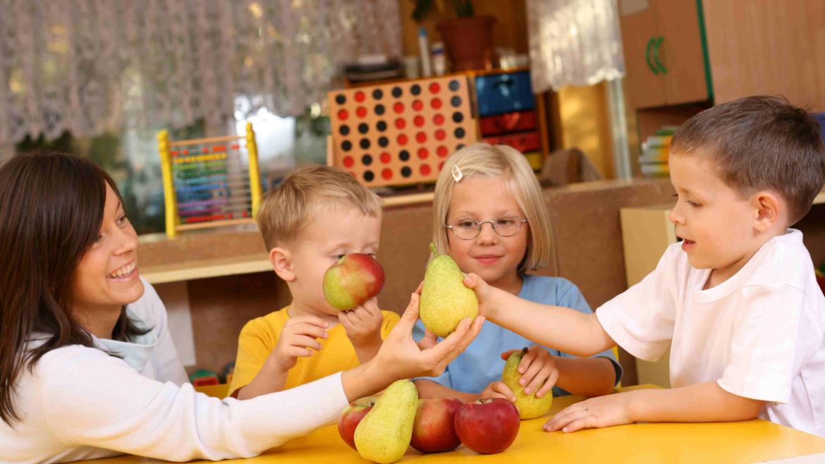 Kinder in der Kita schauen sich mit Erzieherin am Tisch birnen und Äpfel an