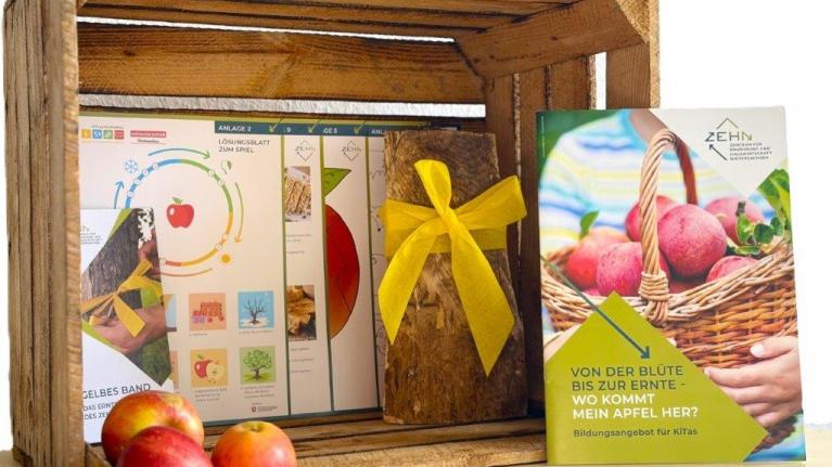 Aufgestellte Holzkiste mit Äpfeln, Broschüre und Glas mit gelbem Band