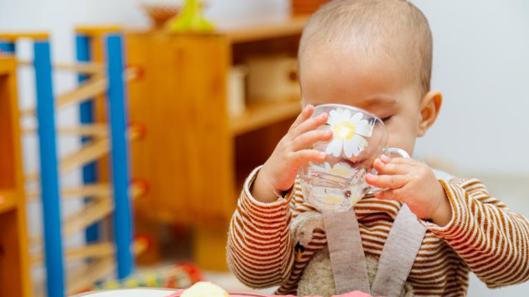 Kleinkind trinkt aus Tasse am Tisch in der Kita