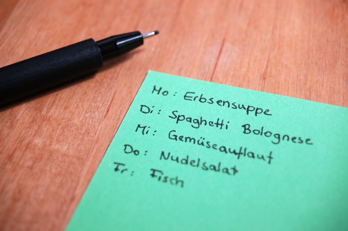 Menüabfolge für die Woche ist per Hand auf einen grünen viereckigen Zettel geschrieben. Montag Erbsensuppe, Dienstag Spaghetti Bolognese, Mittwoch Gemüseauflauf, Donnerstag Nudelsalat, Freitag Fisch