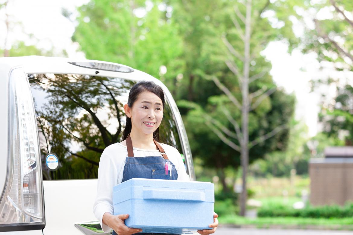 Frau vor Auto mit Warmhaltestyroporbox in der Hand. Die Box ist hellblau.