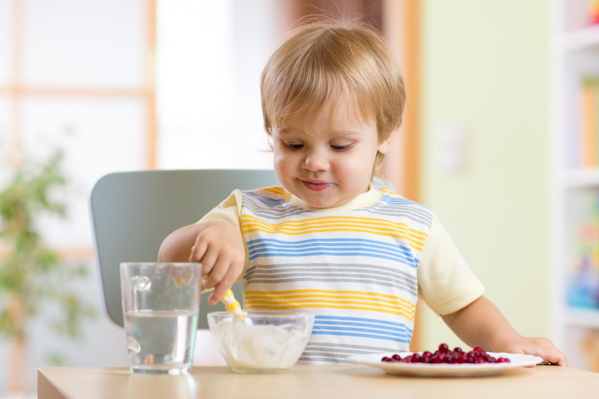 Kind sitzt an einem Tishc und rührt mit dem Löffel in einem Joghurt. Daneben steht ein Teller voller Beeren