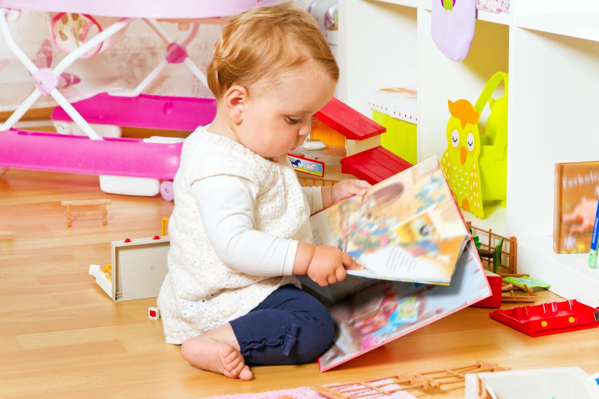 Kleinkind  sitzt auf Boden und schaut ein Bilderbuch an. Im Hintergrund ist ein Puppenhaus.