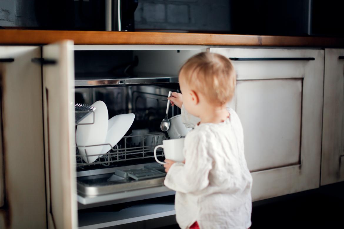 Kleinkind in weißer Kleidung räumt Geschirr in eine Spümaschine.