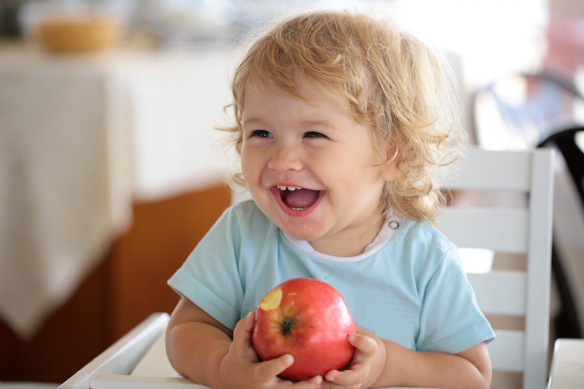 Kind lacht und isst Apfel. Sitzt auf Stuhl in der Kita.