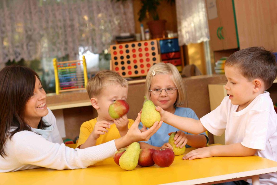 Kinder in der Kita schauen sich mit Erzieherin am Tisch birnen und Äpfel an