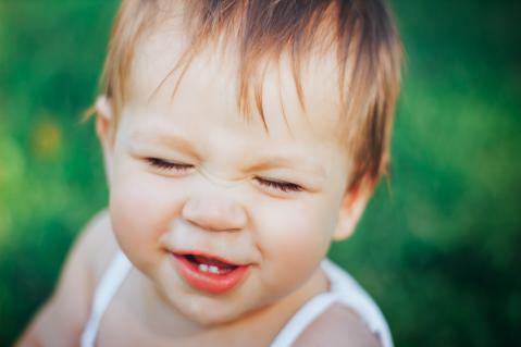 Kind hat Augen zu und den Mund auf, man sieht vorne 2 kleine Zähne. Hintergund ist verschwommen grün.